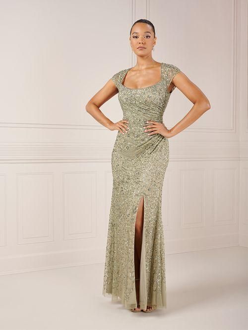 Woman in a beaded, sage green gown with a thigh-high slit, arms akimbo, in a white-walled setting.