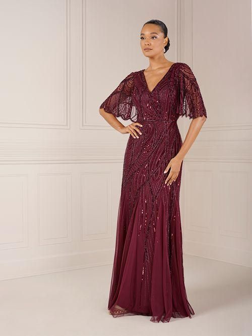 Woman in a long, burgundy gown with beaded bodice and cape sleeves, posing indoors.