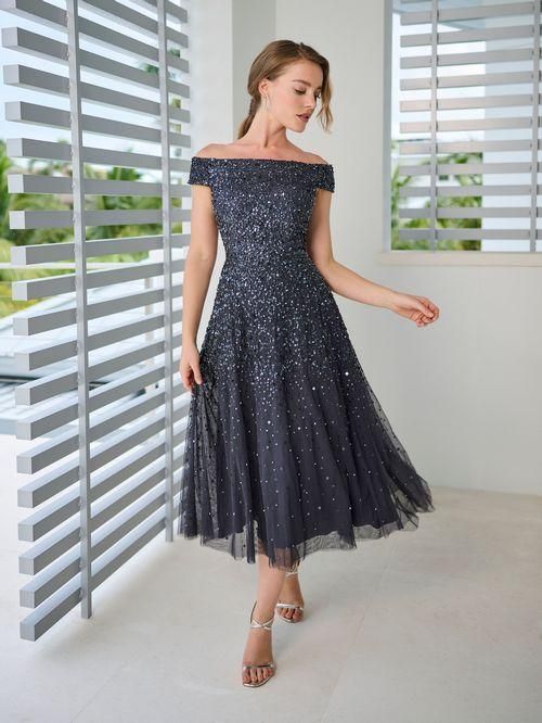 Woman in a dark, sparkling off-shoulder dress, posing by a window with a light, open-air background.