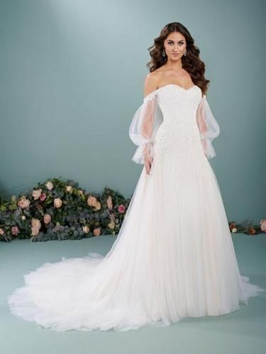 Bride in a white off-the-shoulder wedding dress with long sleeves and a flowing train, standing in front of floral decorations.