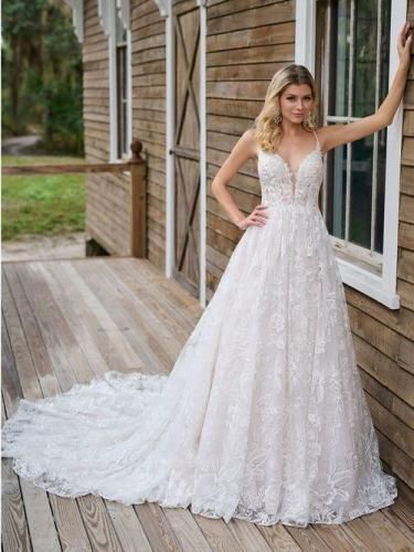 Woman in a white lace wedding dress poses by a wooden building, smiling.