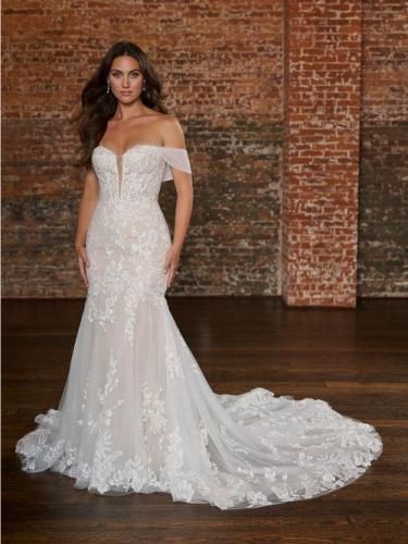 Bride in a white lace off-the-shoulder wedding gown posing in a brick-walled room.