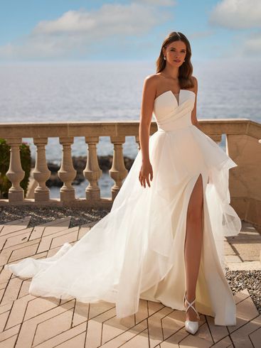 Woman in a strapless white wedding dress with a high slit, standing on a balcony overlooking the ocean.