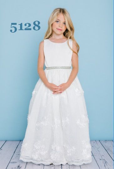 Blonde girl in a white dress with floral details and a jeweled belt, standing in front of a blue background.