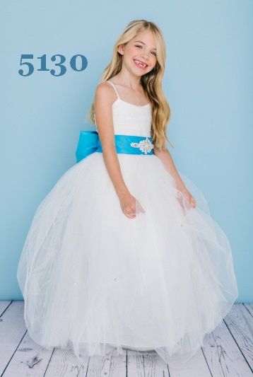 Girl with blonde hair wearing a white ball gown with blue sash, smiling, standing on wooden floor.