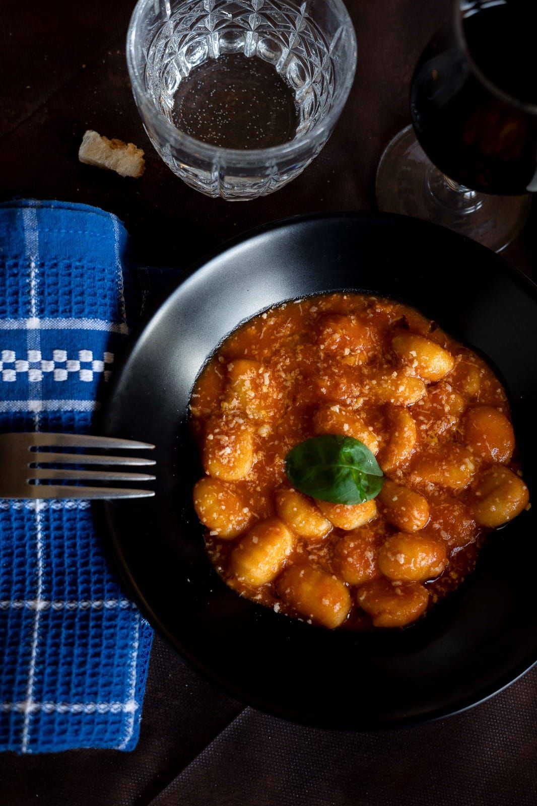 Una ciotola nera di gnocchi in salsa rossa al basilico, accanto a un bicchiere, una bottiglia e un tovagliolo a quadretti blu su un tavolo.