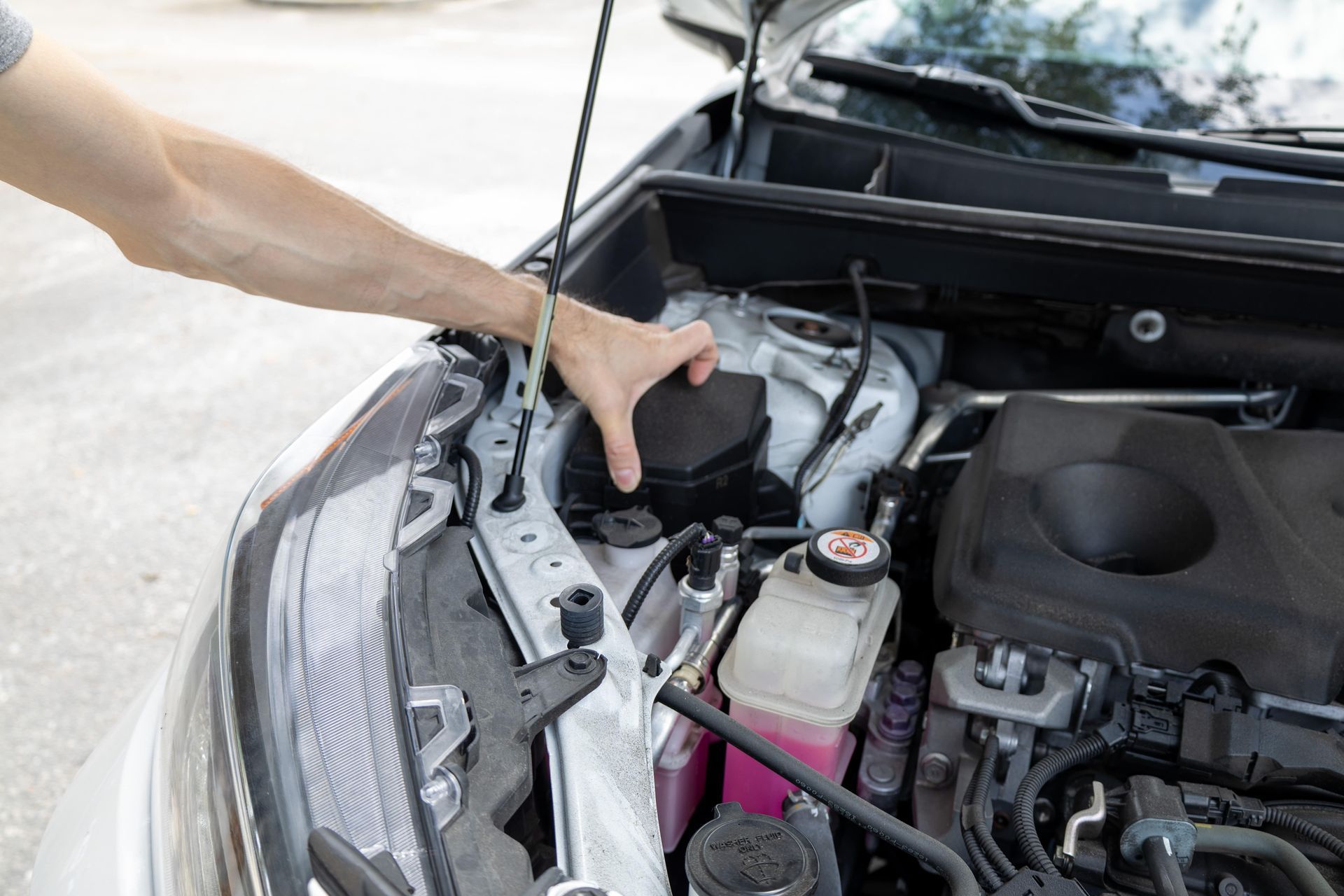 Person reaching into a car engine compartment. Hood is open, and engine components are visible.