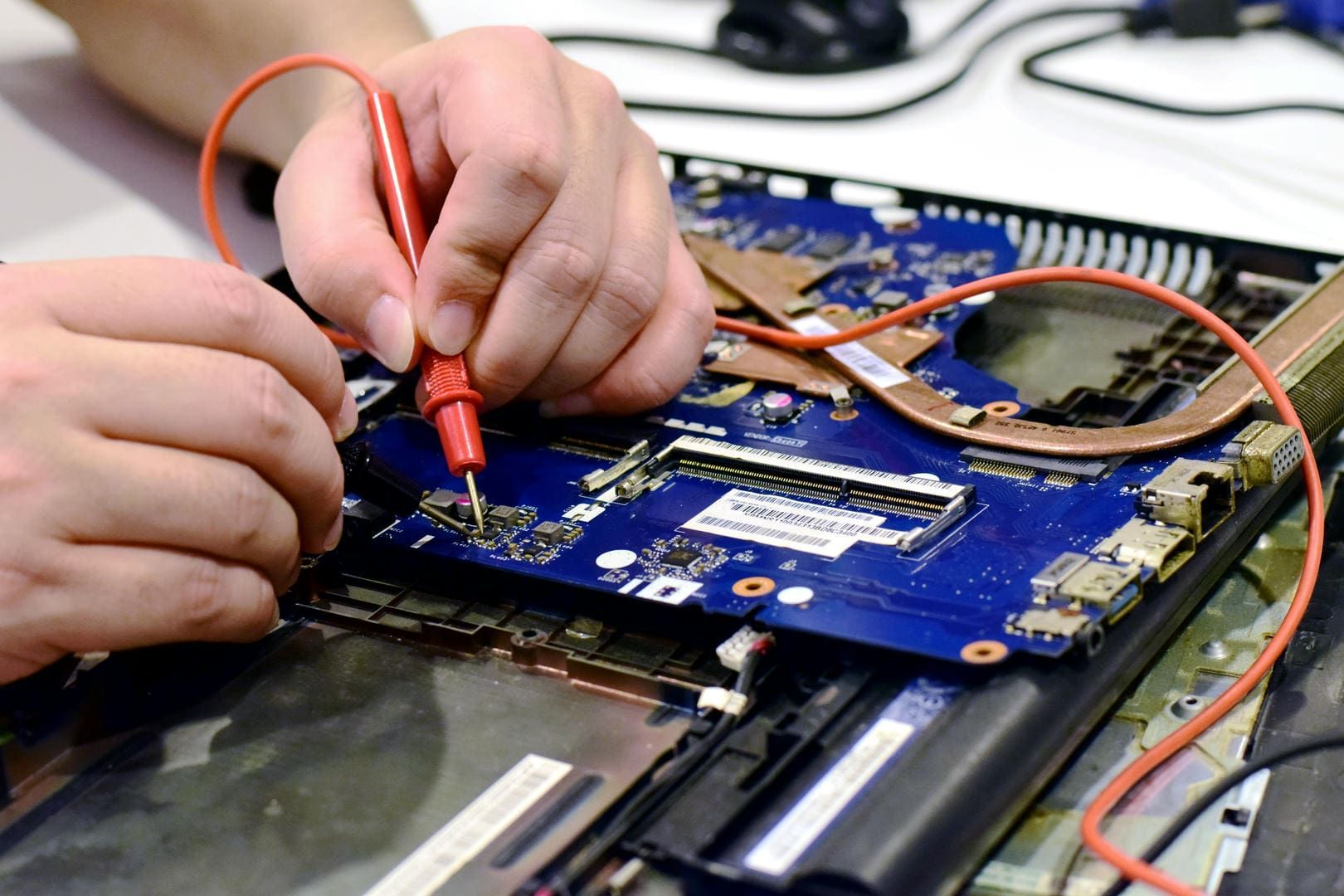 Hands using probes to test a laptop's circuit board, with wires and components visible.
