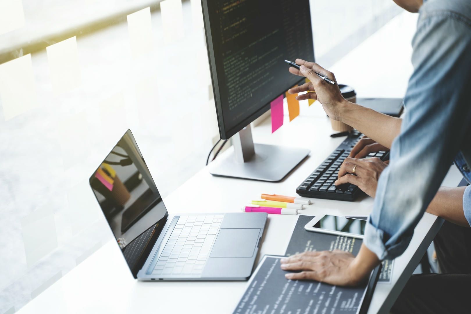 Two people working on computers, one points at the monitor with code, a laptop and documents are on the desk.