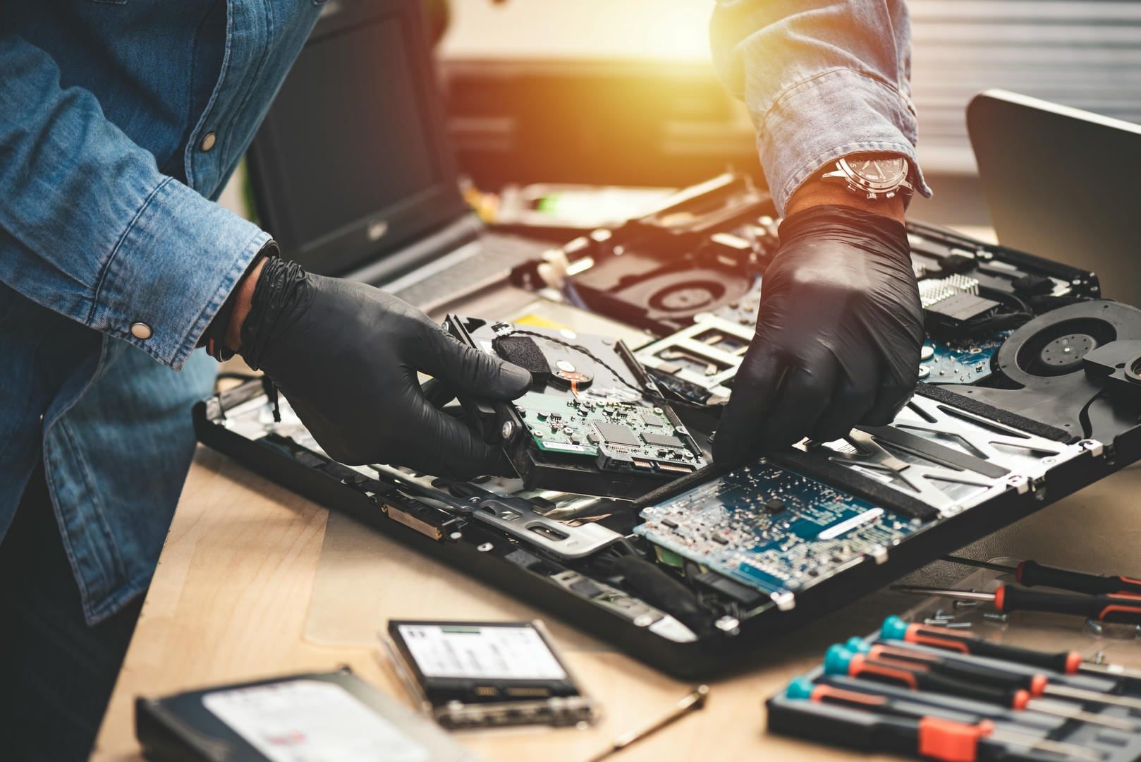 Person in gloves repairs a laptop, holding a circuit board with tools and parts on a table.