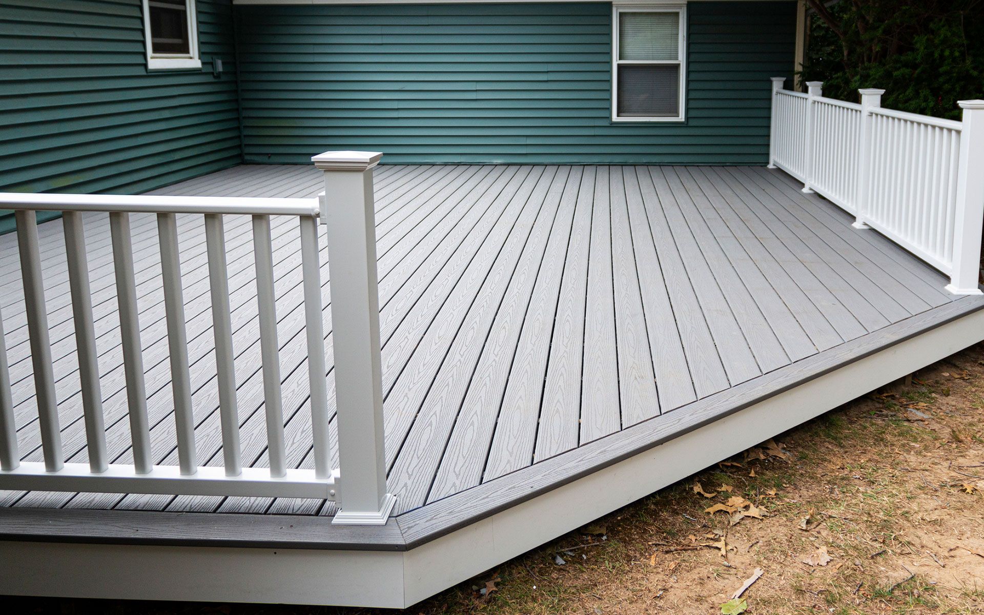 A gray deck with a white railing is in front of a green house.