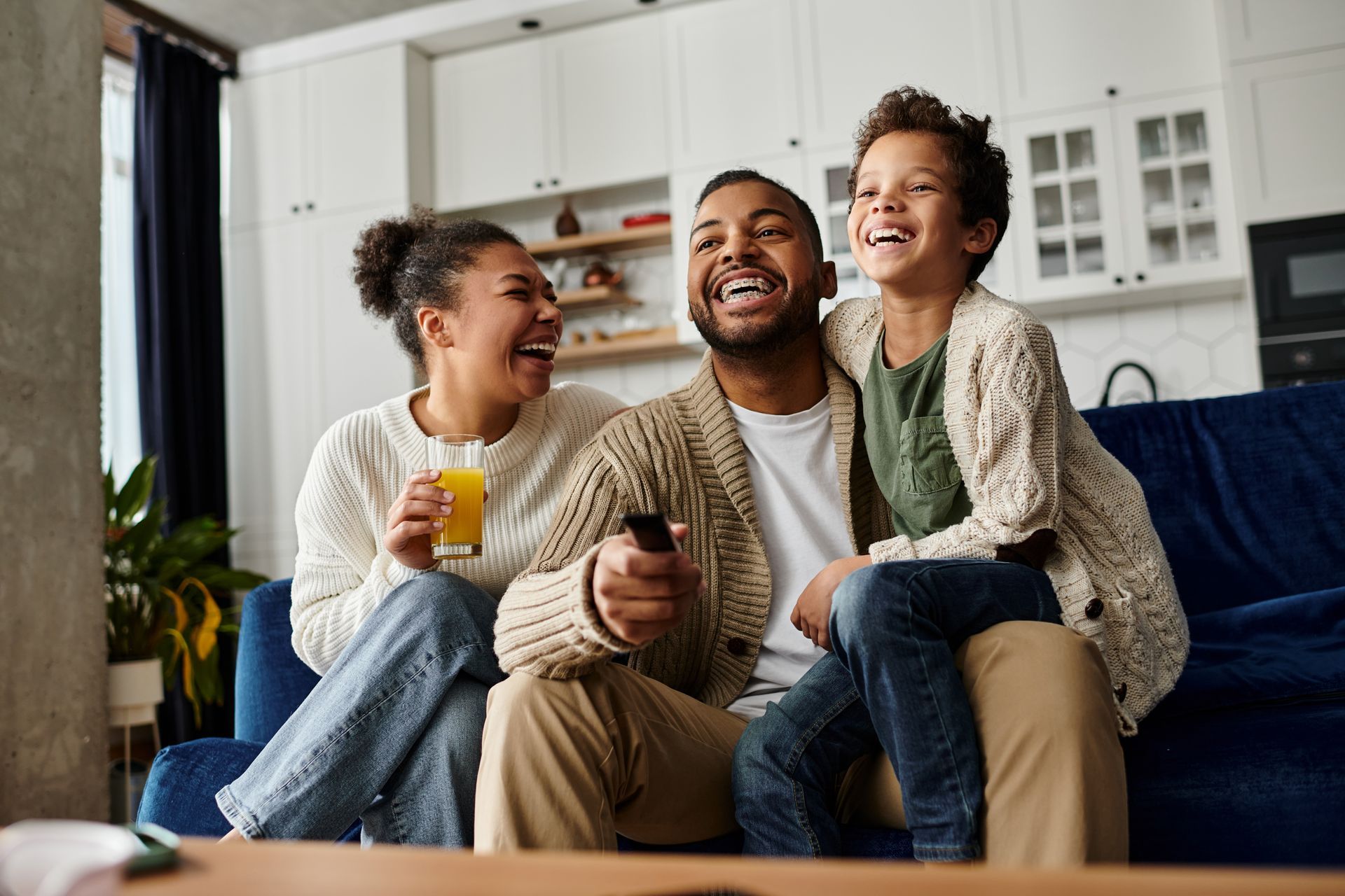 Family laughing while sitting on a couch, holding a remote control and drink. In a home setting.