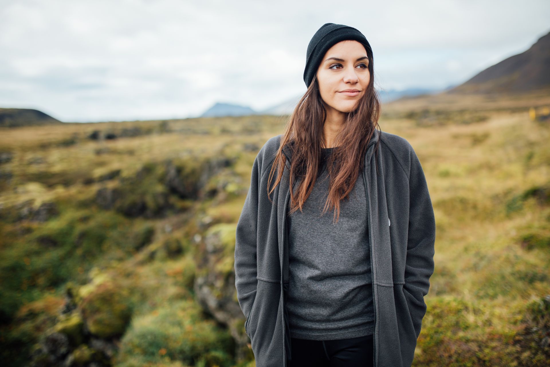 Woman in beanie and coat, standing in a field with mountains in the background, looking off to the side.