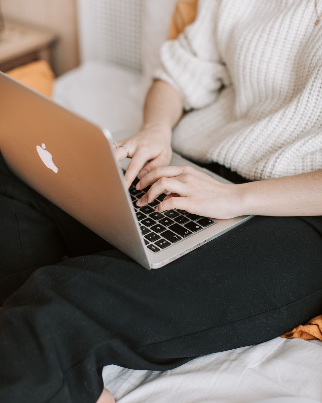 Person typing on a laptop while sitting cross-legged on a bed in a cozy room