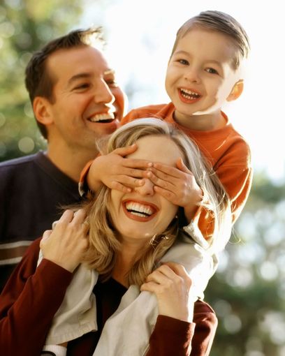 Smiling family outdoors, child covering a woman’s eyes while a man laughs behind them.