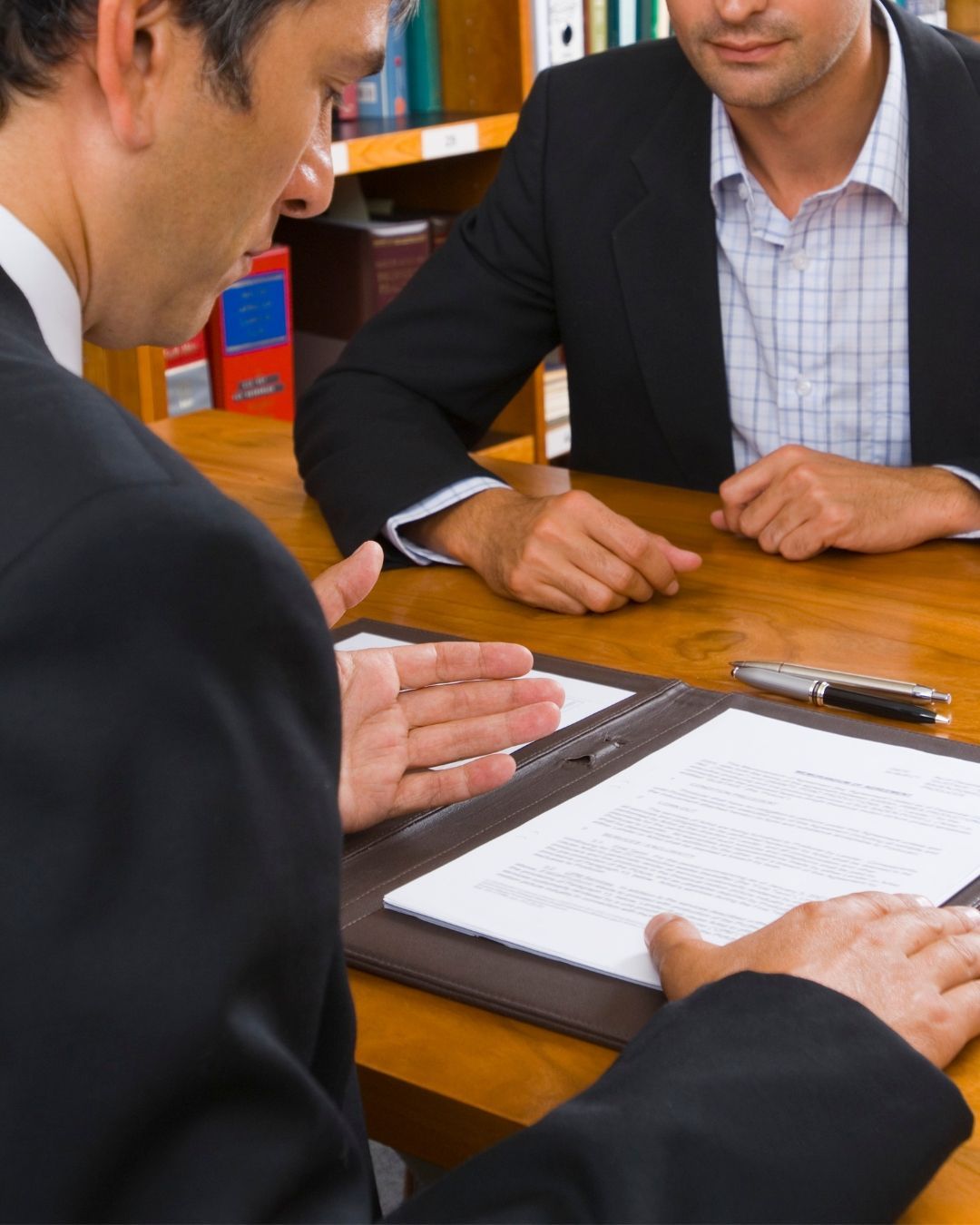 Two people in suits discussing paperwork across a wooden table in an office.