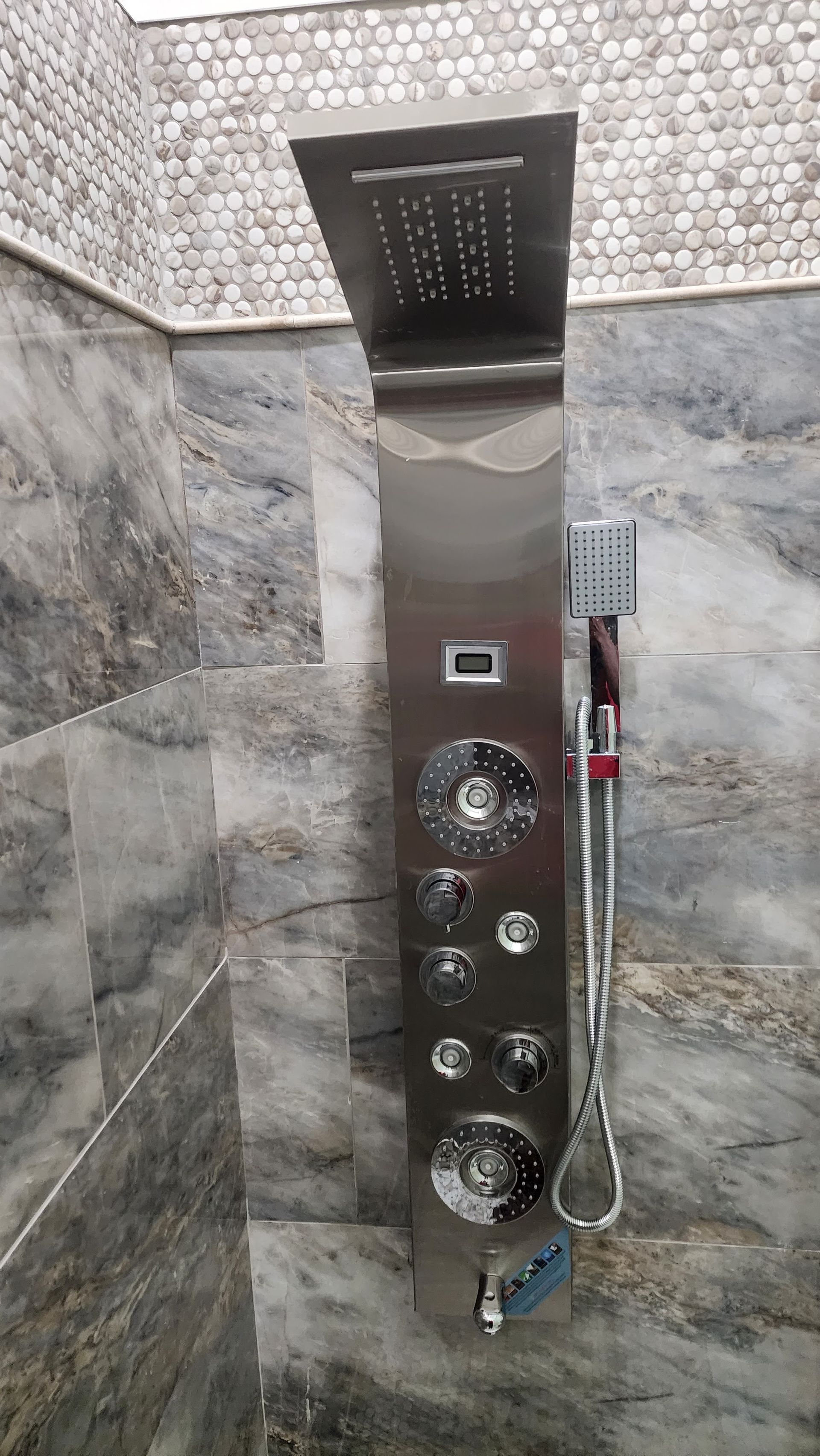 Stainless steel shower panel with various spray heads and handheld shower against marble tile.