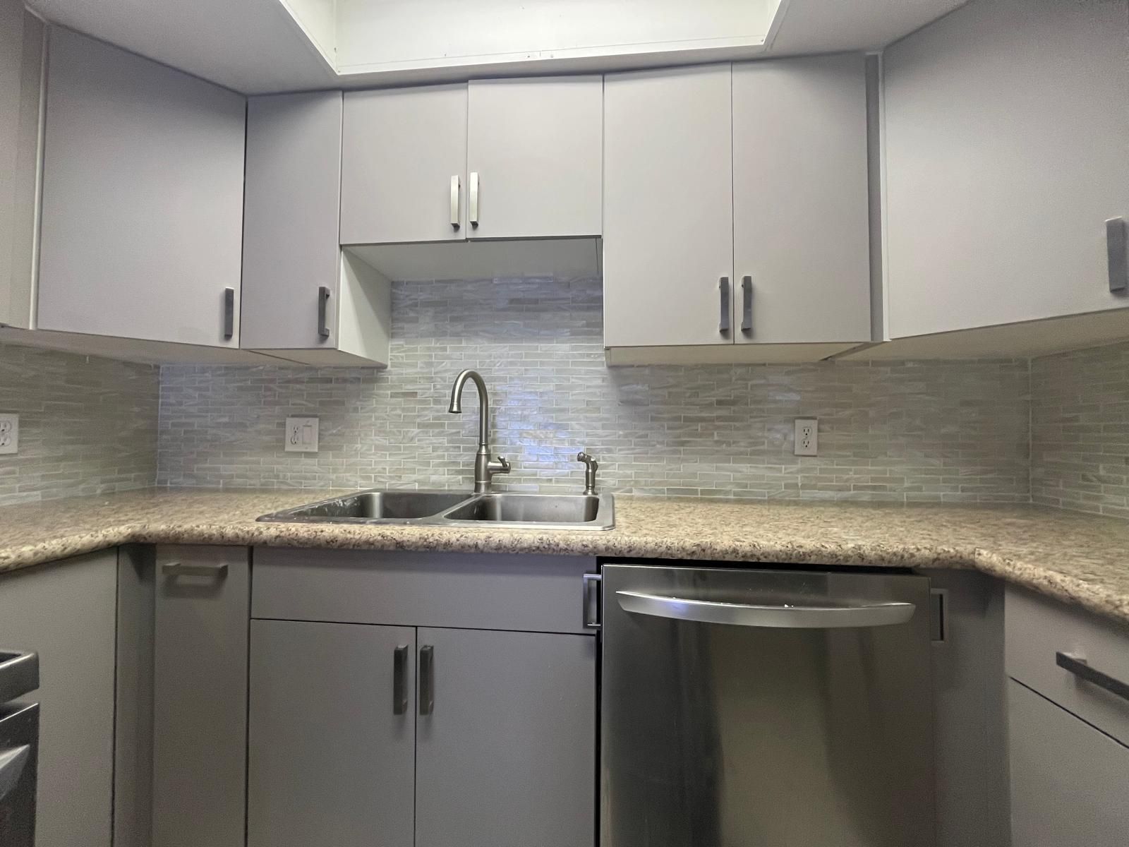 Gray kitchen cabinets with a stainless steel sink and appliances. Light gray backsplash and countertops.