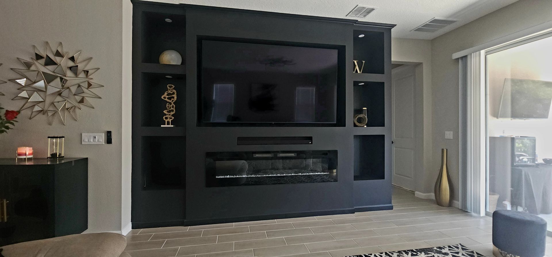 Black built-in entertainment center with a fireplace and large TV. Decorative objects and sliding glass door on the right.