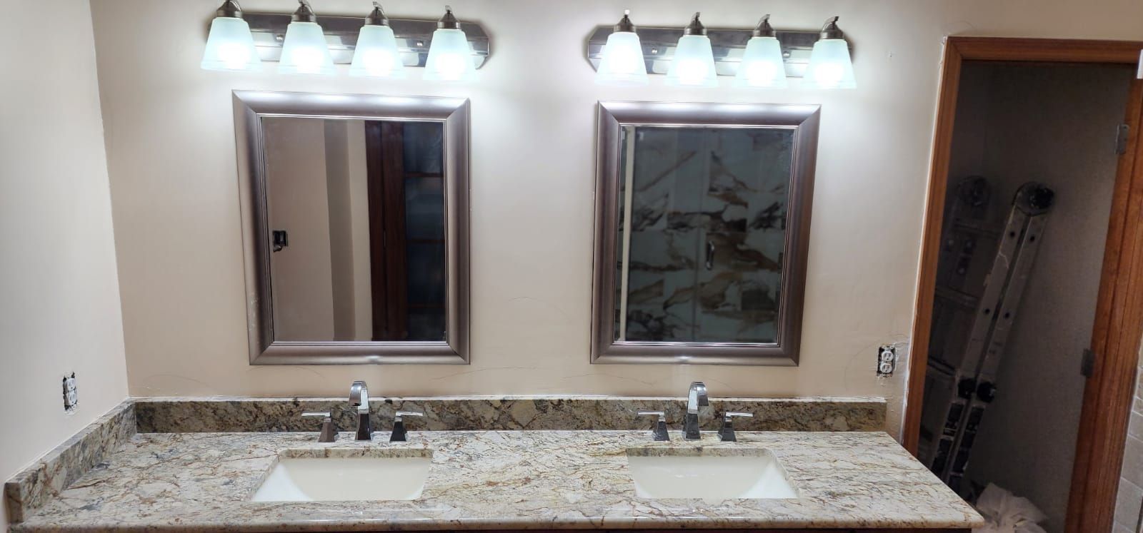 Bathroom with two sinks, mirrors, and lights above a speckled countertop. A door with a ladder is on the right.