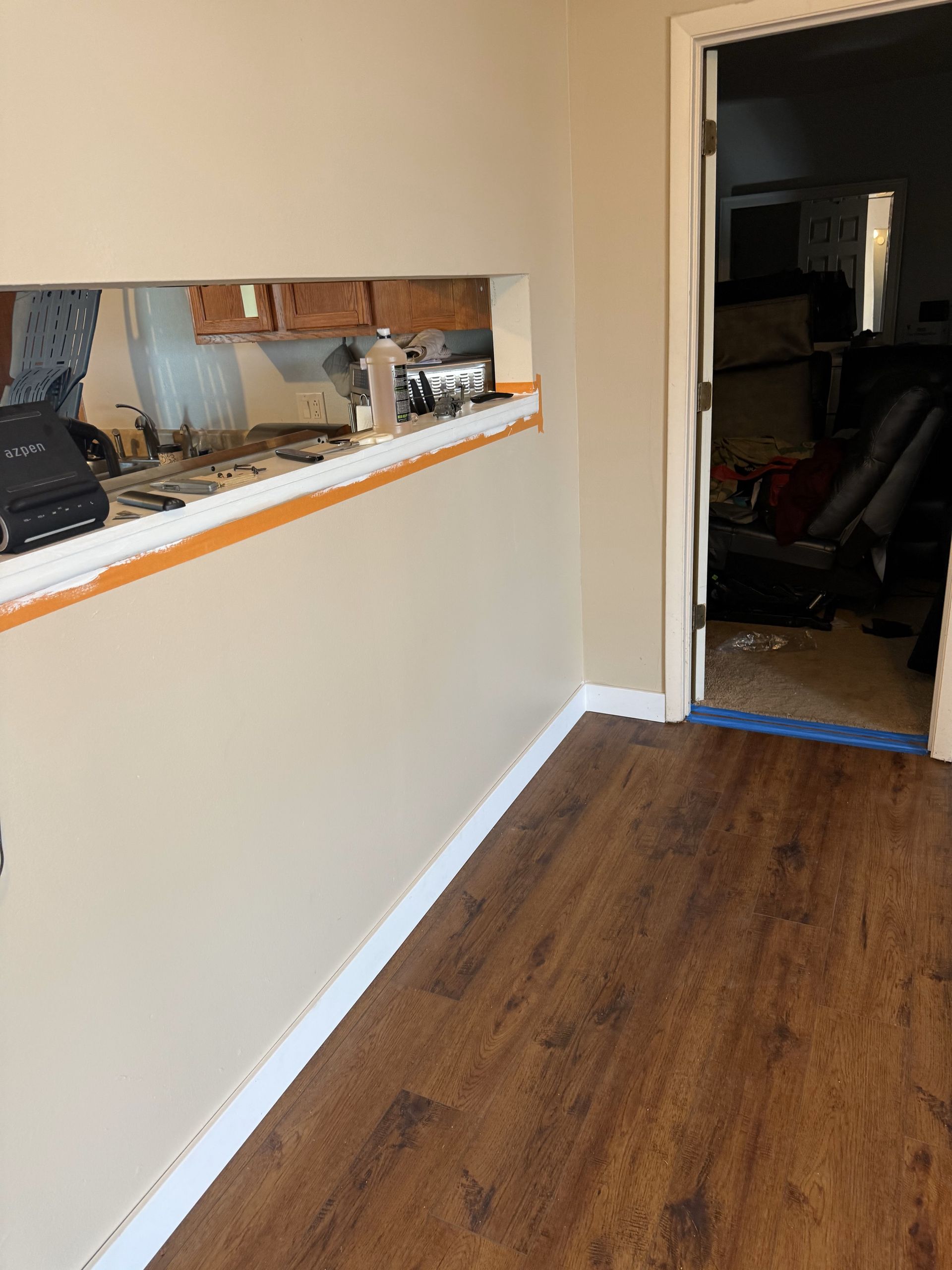Room interior with an opening to a kitchen, brown floor, tan walls, and white trim.