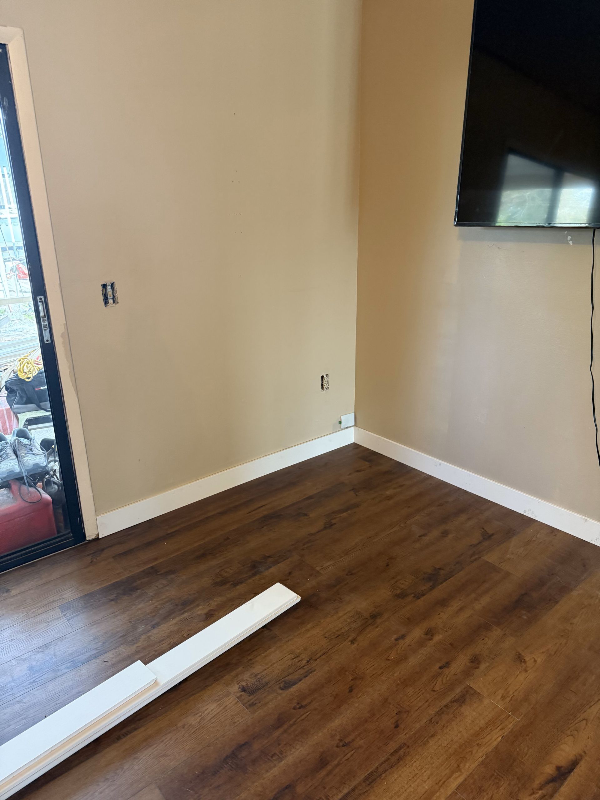 Empty room with wood floors and tan walls. Sliding glass door and TV on the wall. Trim is in process.