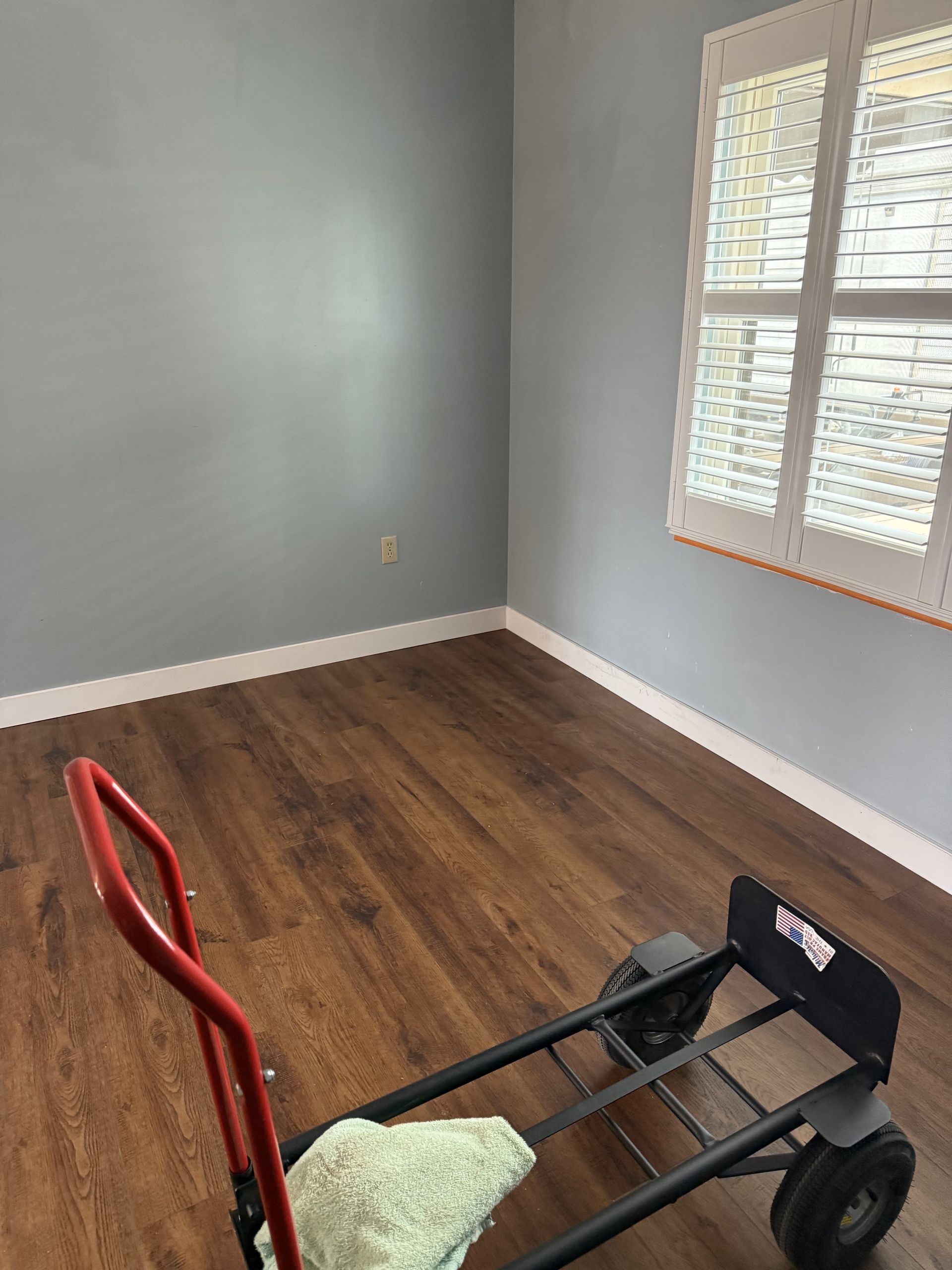 Empty room with blue walls, wood-look flooring, and a window with blinds. A hand truck rests in the foreground.