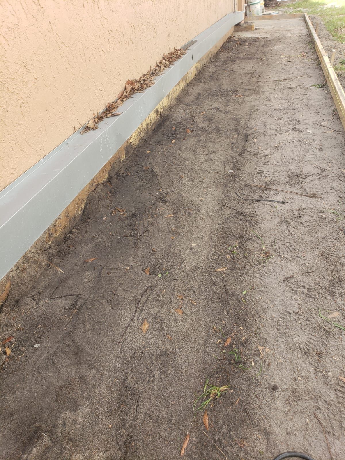 Narrow dirt pathway alongside a beige building with a grey gutter. Wooden border on one side, debris present.