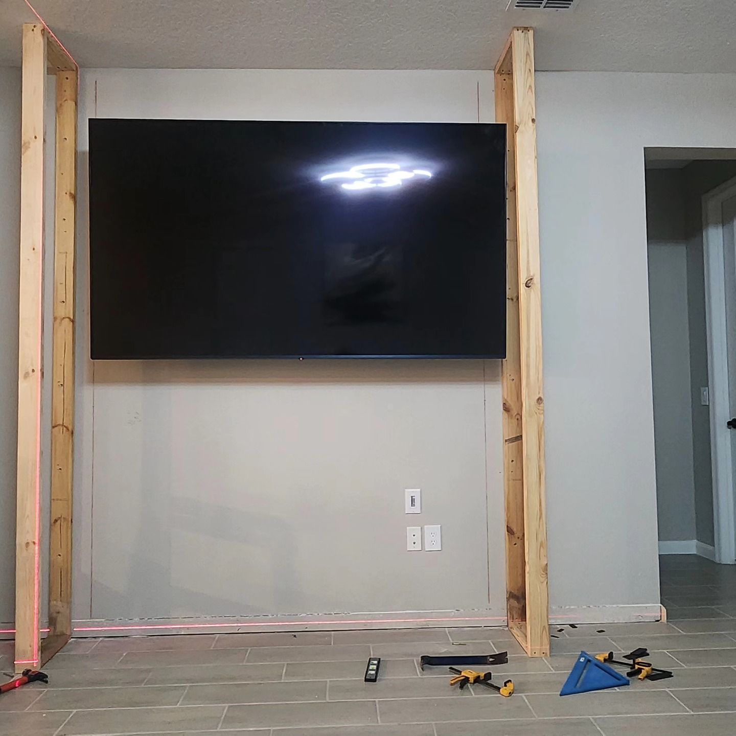 TV mounted on a wall within a wooden frame, tools on the floor.