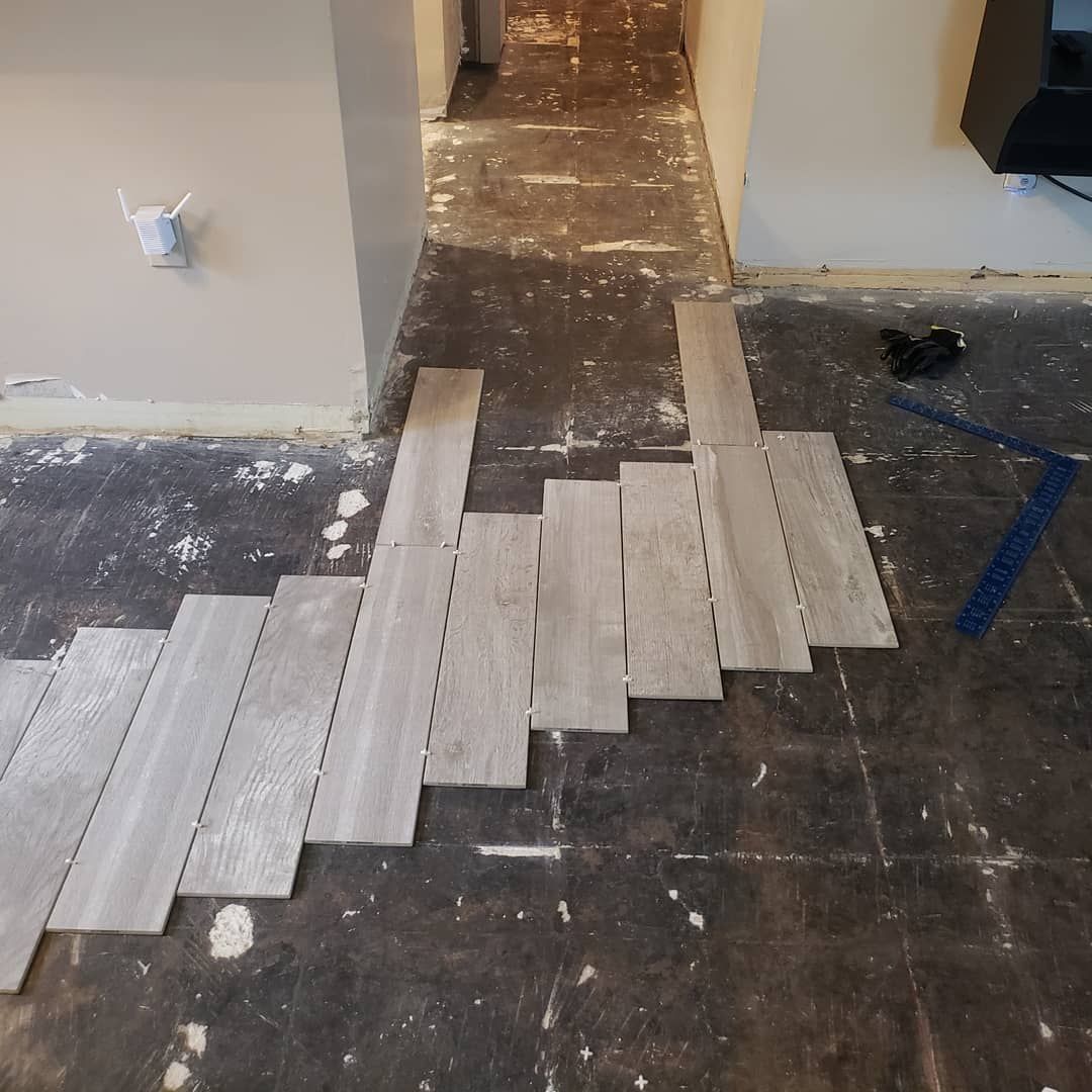 Wooden-look tiles being laid on a dark floor in a room. The floor shows signs of previous flooring removal.