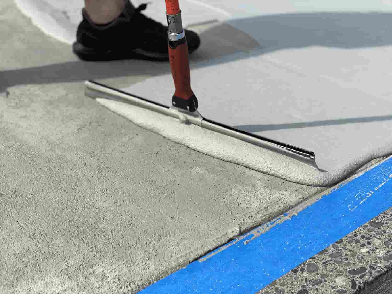 Person using a squeegee to spread gray coating on a concrete surface, near blue tape.
