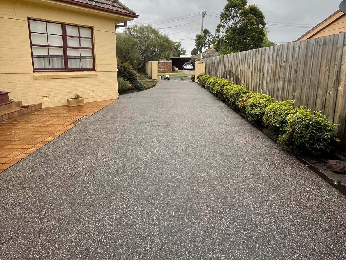 A gravel driveway extends from the street toward a house, bordered by hedges and a wooden fence.