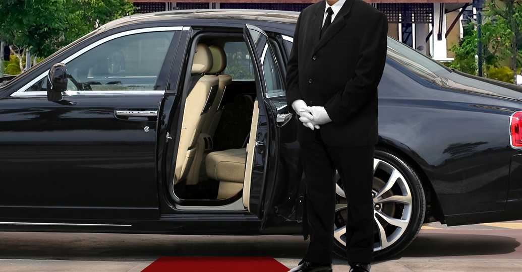 Butler in black suit opening car door for passenger with red carpet.