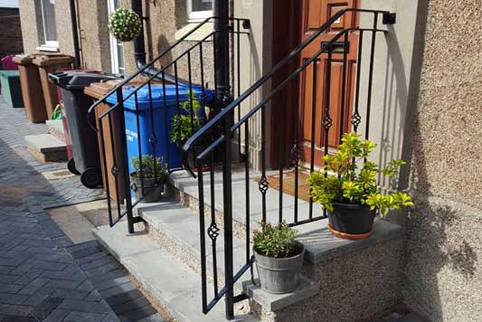 Bespoke handrails with flagged steps, a brown door, potted plants and blue, brown and black wheelie bins