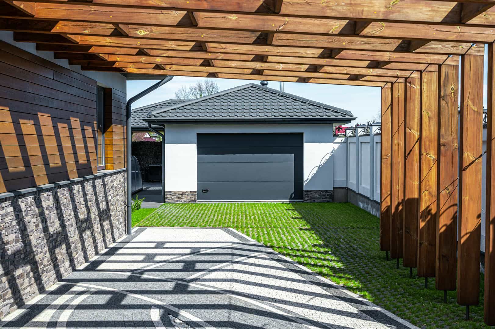 Wooden pergola framing a driveway leading to a garage with a black door and a grass-covered driveway.