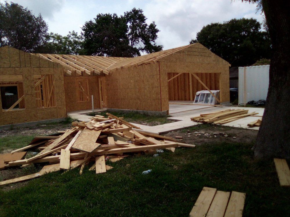 House under construction: wooden frame with open garage, debris pile in front, cloudy sky.