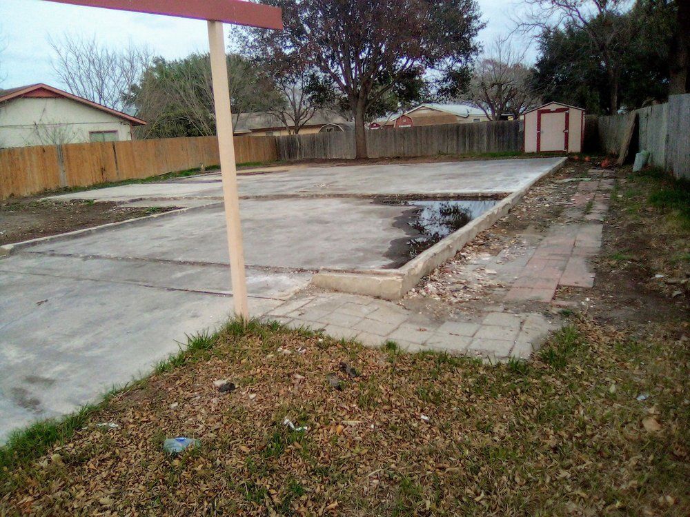 Concrete lot with a brown pole, surrounded by a wooden fence, grass, and a shed.