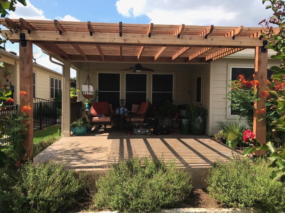 Wooden pergola over a patio with seating, plants, and a fan. Sunny outdoor setting.