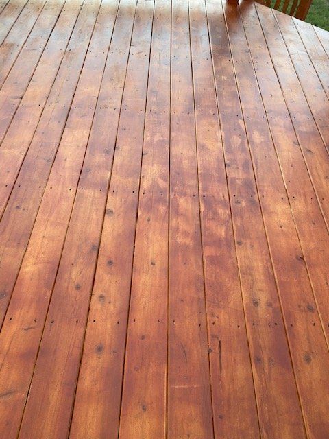 Wooden deck, stained reddish-brown, viewed from above. Boards run lengthwise, with gaps between them. Sunny outdoor setting.
