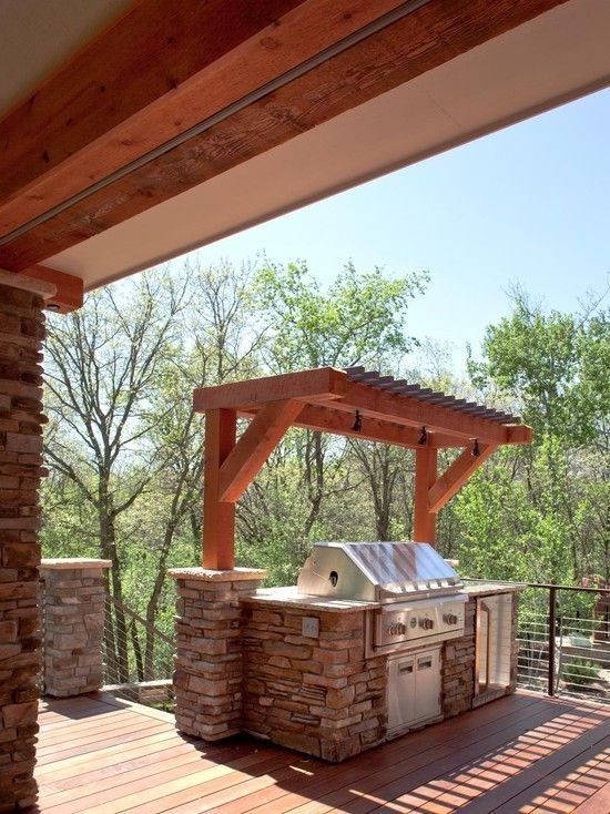 Outdoor kitchen with stone base, wooden beams, stainless steel grill, and a forest backdrop.
