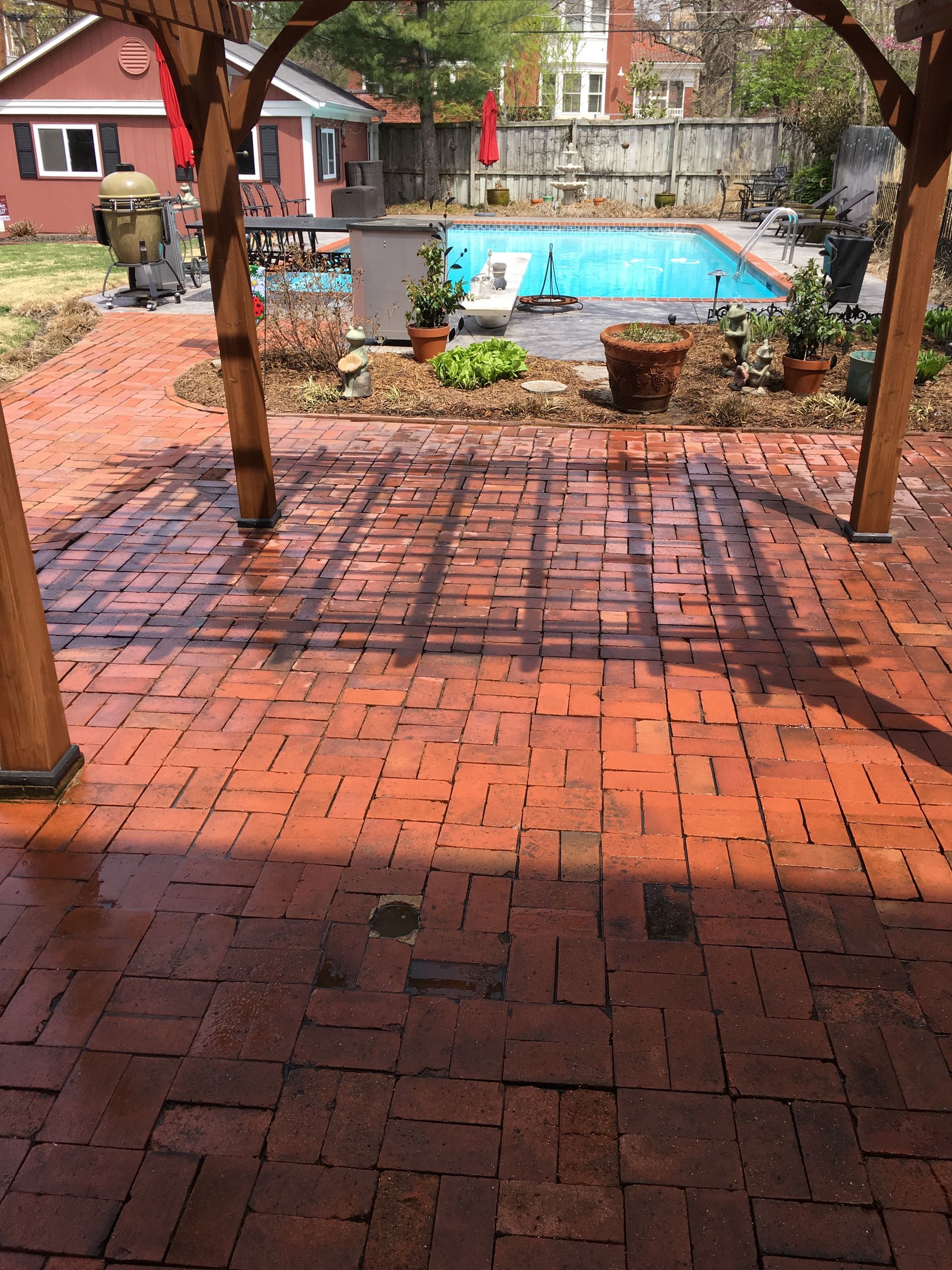 Red brick patio with a wooden pergola, leading to a blue swimming pool in a backyard.