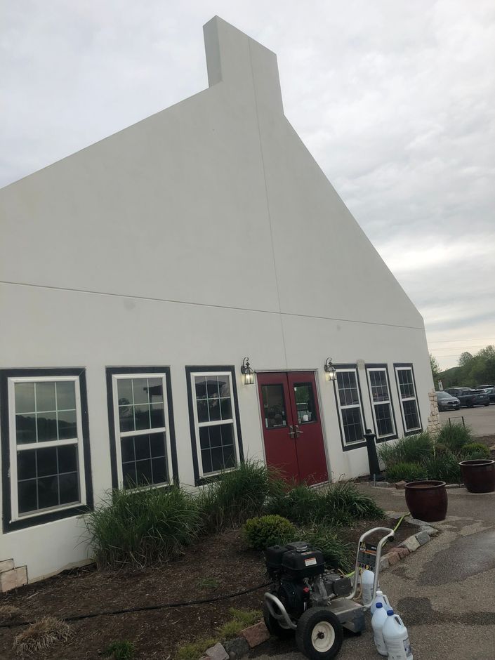 White building with black-trimmed windows, red door, and landscaping, with a pressure washer in front.