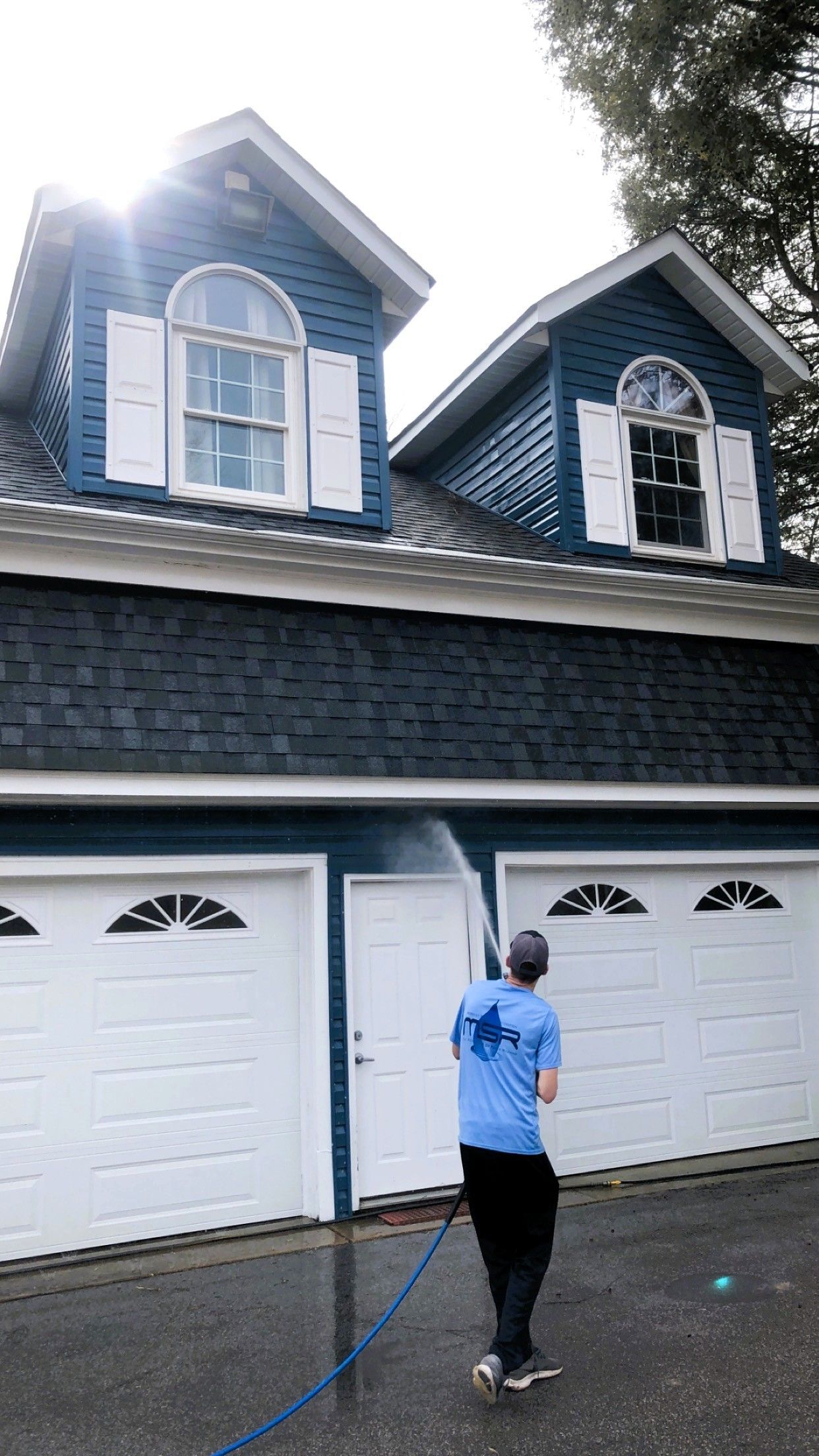 Man spraying water upwards near a blue-sided house with white garage doors.