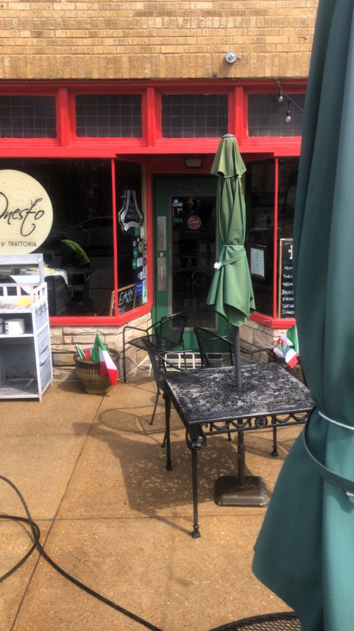 Exterior view of an Italian cafe with outdoor seating and green umbrellas. Red trim, brick building.