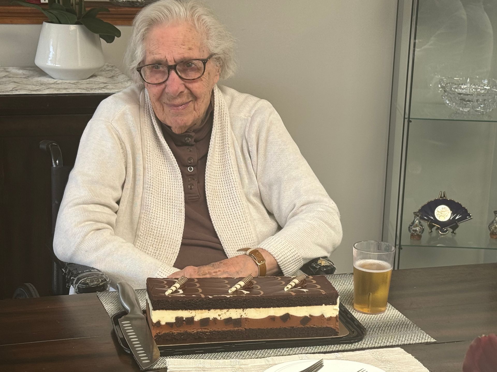 An elderly woman is sitting at a table with a cake and a glass of beer.