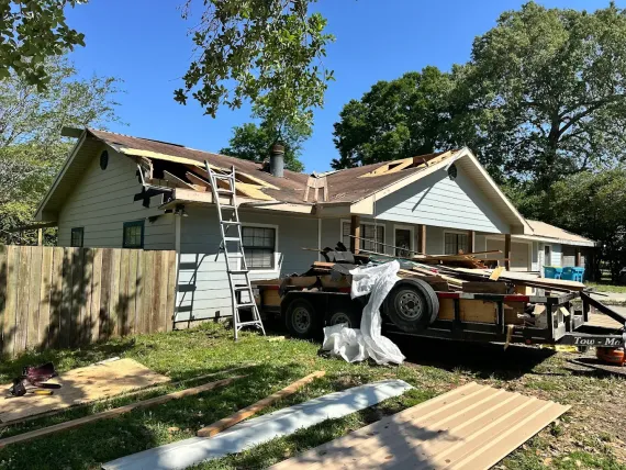 House with partially removed roof; trailer with debris; ladder leaning against the house; construction site.