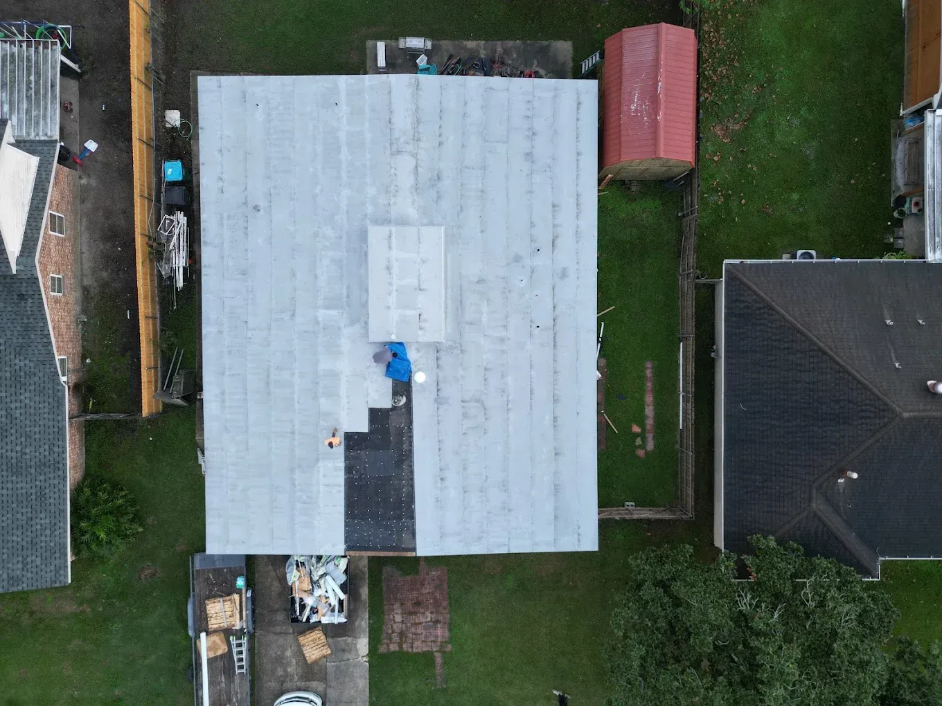 Aerial view of a house with flat roof under repair; a person is working on the roof, surrounded by materials and a storage shed.