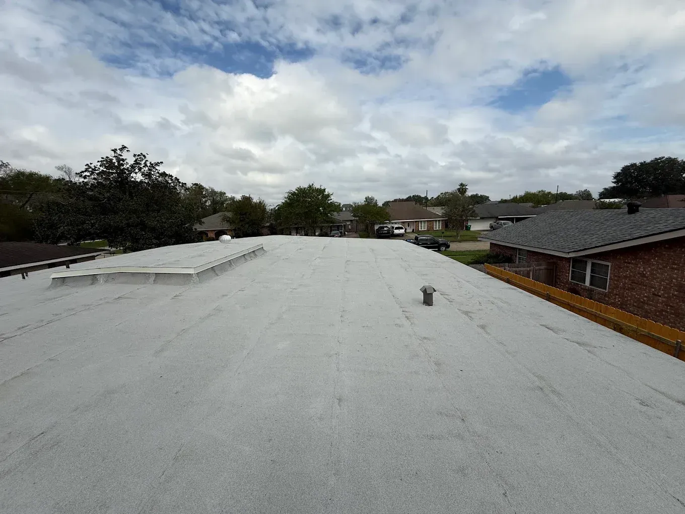 A flat, light-colored roof with some vents under a cloudy sky; view of a neighborhood.