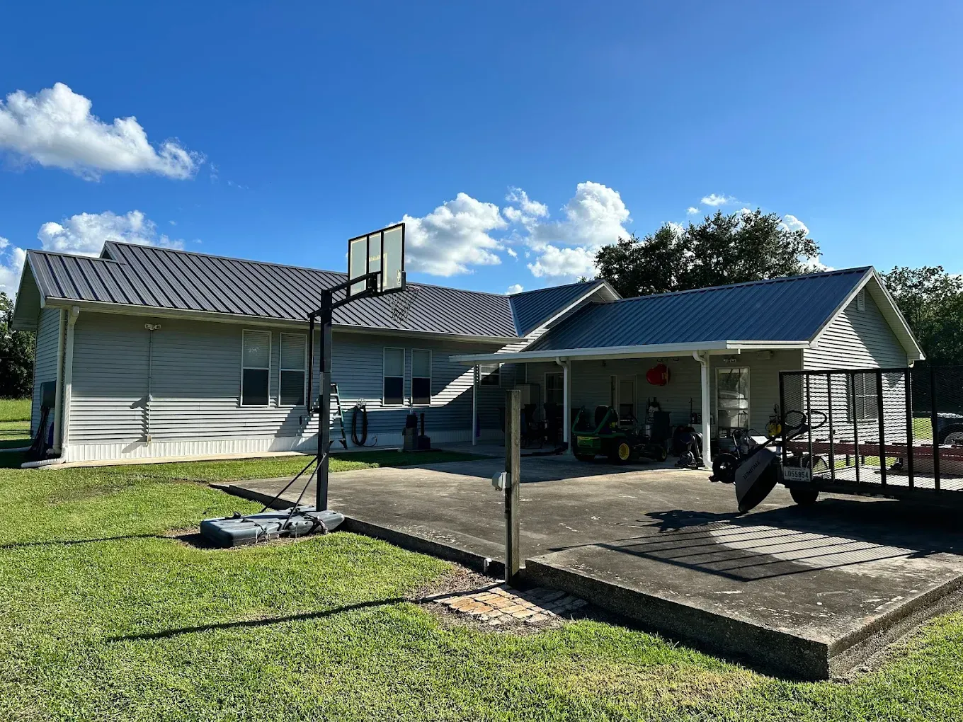 Exterior view of a house with metal roof, basketball hoop, and trailer on a sunny day.