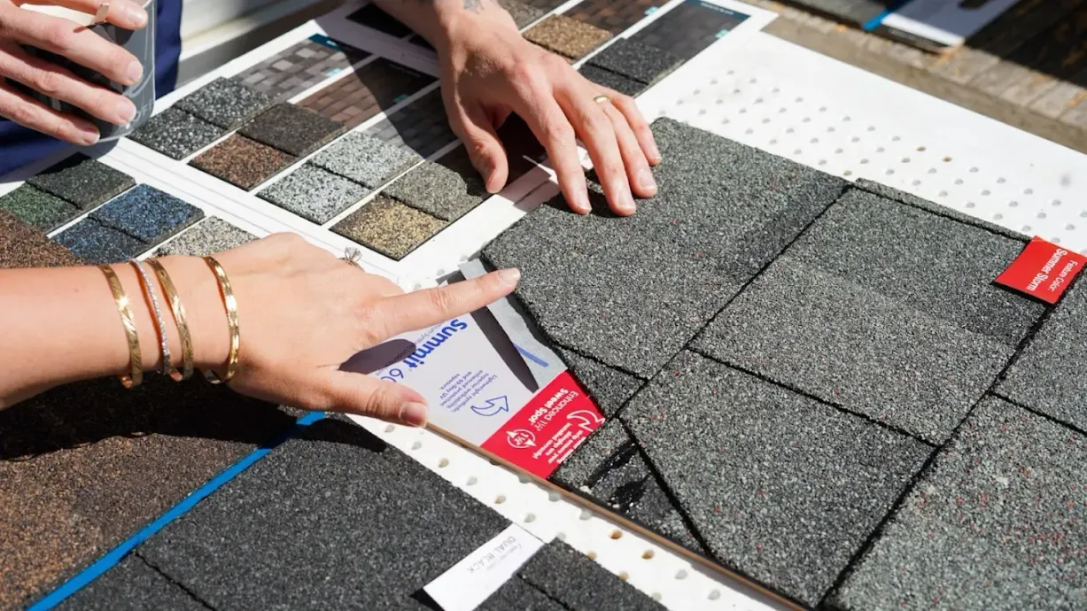 Hands pointing to gray roofing shingles and color samples on a table.